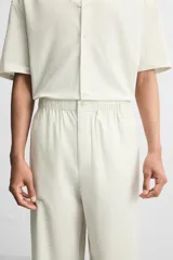 Pantalón relaxed fit de color beige con finas rayas verticales, confeccionado en una mezcla de algodón y cáñamo. Presenta cintura elástica, bolsillos laterales, un bolsillo trasero de plastrón y cierre frontal con botones.