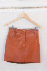 Minifalda de cuero color camel, de corte recto, con cierre frontal y botones visibles en la cintura. Presenta bolsillos delanteros tipo parche.
