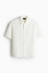Camisa blanca de punto con textura acanalada, corte regular, cuello inglés, tapeta lisa, canesú en la espalda y bajo recto con aberturas laterales.