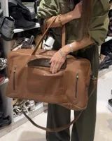 Bolso de viaje tipo weekender de cuero vacuno color marrón, con doble asa de mano y correa bandolera desmontable. Posee un bolsillo externo con botón a presión y dos bolsillos laterales con cierre. Producto de fabricación artesanal.