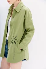 Campera recta color verde claro confeccionada en gabardina, con cuello camisero, mangas largas y bolsillos ojal. Presenta bordados calados en forma de palmeras en el frente y la espalda.