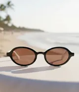 Lentes de sol con montura ovalada de acetato negro y lentes color marrón. Presentan un detalle en la parte superior de las patillas en color blanco.