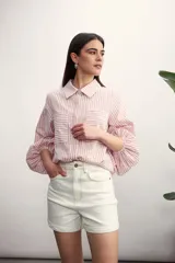 Camisa a rayas verticales color beige y blanco, con cuello clásico, cierre frontal con botón dorado, mangas largas abullonadas con puños elásticos, dos bolsillos frontales y corte holgado.