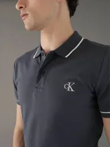Chomba Calvin Klein color gris con cuello polo, tapeta de dos botones, logo bordado en el pecho y ribetes blancos en cuello y mangas. Corte slim fit.