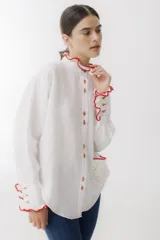Camisa blanca de lino con cuello mao, manga larga abullonada y puños con detalle de crochet rojo. Tiene bolsillo de crochet en el pecho y cierre frontal con botones de madera.
