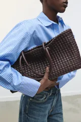 Bolso shopper alargado color marrón chocolate con diseño trenzado. Tiene doble asa de hombro y cierre superior con botón imantado. Incluye una bolsa interior con cierre fruncido.