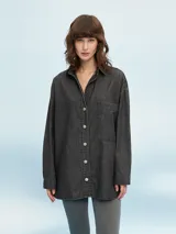 Camisa de denim negro con corte oversize, cuello clásico, cierre frontal con botones metálicos y bolsillo de parche en el pecho.
