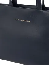 Cartera tipo tote de cuero sintético granulado color azul marino, con dos asas superiores y correa cruzada ajustable con cinta tejida a rayas azules y rojas. Presenta un cierre de cremallera principal y logo metálico de Tommy Hilfiger en el frente.