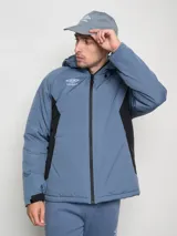 Campera corta de abrigo Umbro Crafted Pro, color negro con paneles laterales en gris. Presenta capucha con cordón ajustable, cierre frontal completo y logo de la marca bordado en el pecho.