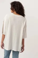 Remera de algodón color blanco hueso, con calce relajado y hombros caídos. Presenta un estampado central con la palabra "HOWDY" en negro y múltiples ilustraciones de botas vaqueras en tonos azules, naranjas y amarillos.