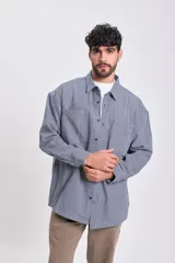 Sobrecamisa de hombre de jean, color azul, con patrón de rayas verticales finas. Presenta cuello camisero, bolsillos frontales con tapa y botones oscuros. Se lleva abierta sobre una remera blanca.