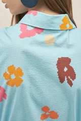 Vestido camisero celeste con estampado de flores de colores, confeccionado en tejido seersucker de algodón con elastano. Tiene cuello camisero, botones nacarados, lazo para ajustar la cintura con terminación en cobre y bolsillos laterales.