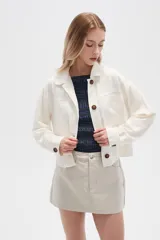 Chaqueta color blanco hueso de lino con cuello camisero, cierre frontal con botones marrones, bolsillos de parche en el pecho y corte holgado.