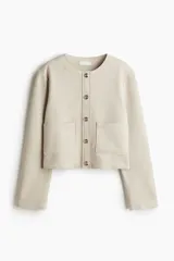 Saco corto color beige de tejido de punto con acabado cepillado suave, cuello redondo, cierre frontal con botones metálicos dorados y bolsillos de parche delanteros.