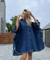 Camisola tipo poncho en jean azul oscuro con cuello camisero, cierre frontal con botones, mangas cortas amplias con volados y ruedo con flecos.
