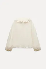 Blusa color crudo confeccionada en hilatura con mezcla de lyocell. Escote pico ajustable con cordones, manga larga con volumen acabada en elástico y detalle de volantes en el mismo tejido.