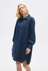 Vestido camisero de jean azul oscuro, corte holgado, largo a la rodilla, con cuello camisero, mangas largas abullonadas con puños abotonados y cierre frontal con botones.