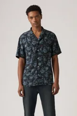 Camisa de hombre de manga corta con estampado floral en tonos azules sobre fondo negro. Tiene cuello tipo cubano, cierre frontal con botones y un pequeño logo rojo bordado en el pecho.