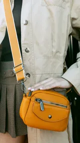 Bandolera naranja de cuero sintético con bolsillo frontal con cierre y bolsillo pequeño con tapa y broche magnético. Cuenta con apertura central superior con cierre, bolsillos internos y correa larga regulable y desmontable.