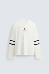 Camiseta blanca de manga larga con cuello en pico. Tiene el número 12 bordado en el pecho y dos franjas horizontales en color azul marino en cada manga.