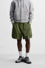 Bermuda color verde militar, relaxed fit, confeccionada en tejido técnico ligero. Cintura elástica ajustable con cordón interior, pliegues frontales, bolsillos delanteros y traseros con solapa, bolsillos cargo con solapa en perneras. Cierre frontal con cremallera y botón.