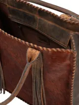 Bolso tipo shopper de piel con pelo en tonos marrones, bordes cosidos a mano con hilo grueso y dos asas de cuero trenzado con flecos largos colgantes.