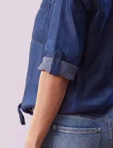 Blusa de tencel azul con cuello a la base y escote en V, abertura central con botones, bolsillos delanteros, mangas 3/4 regulables con presilla y botón, y terminación de ruedo con nudo.