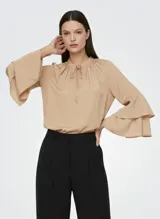 Blusa color beige claro con cuello alto fruncido y lazo ajustable en el escote. Mangas largas con volantes escalonados en los puños. Corte holgado y tejido fluido.