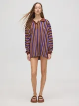 Chomba de corte oversize con cuello camisero, a rayas verticales en color marrón, lila y blanco. Tiene un logo bordado en el pecho y puños en color marrón.