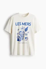 Remera color crudo de algodón con cuello redondo y mangas cortas. Estampado frontal con la frase "LES MERS" y dibujos de conchas y estrellas de mar en color azul.
