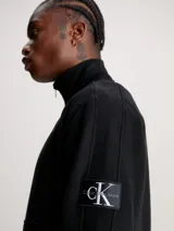 Buzo negro de felpa de algodón orgánico con cuello alto con cremallera, puños y bajo de canalé, bolsillo canguro y parche con logo en la manga.