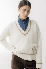 Sweater beige con cuello en V y mangas largas con diseño trenzado. Tiene dos bolsillos frontales con bordados de tréboles rojos y ribetes rojos en cuello, puños y ruedo.