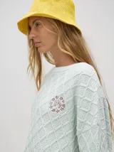 Sweater color crema con tejido en ochos, cuello redondo, puños acanalados con doble raya azul y tajos a los costados.