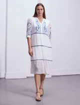 Vestido midi blanco con bordados florales azules en el escote, mangas y cintura. Tiene cuello en V, mangas 3/4 con botón y falda escalonada con detalles de ribetes azules.