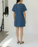 Vestido corto de denim azul, con cuello redondo y mangas cortas amplias. Presenta un corte ligeramente entallado y un cinturón de cadena metálica con eslabones circulares.
