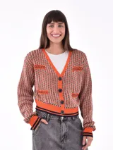 Cárdigan corto de punto con estampado geométrico en tonos naranja, blanco y negro. Tiene cuello en V, cierre frontal con botones negros, dos bolsillos delanteros y ribetes en contraste.