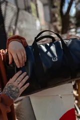 Bolso duffel negro de cuero sintético con logo de ancla bordado en rosa. Tiene doble asa de mano reforzada, correa de hombro regulable y desmontable, cierre superior con doble cremallera y tres bolsillos (dos exteriores y uno interior).