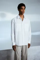 Camisa blanca de popelina de algodón con cuello inglés, tapeta clásica, dos bolsillos superiores abiertos, canesú con pliegue de caja en la espalda, mangas largas con puños abotonados, tapeta de manga con botón y bajo levemente redondeado. Corte holgado.