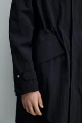 Parka negra de tejido técnico ligero, corte regular, cuello alto con capucha ajustable, manga larga con puños ajustables, cintura ajustable con cordón interno, bolsillos de solapa en la cadera, abertura en la espalda y cierre frontal con cremallera oculta y botones a presión.