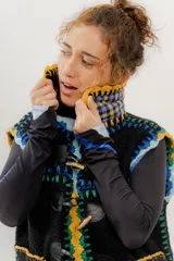 Chaleco negro de corderito con cierre de alamares, tejido a mano en crochet con detalles en amarillo, verde y azul. Cuello alto con interior a cuadros. Talle oversized.