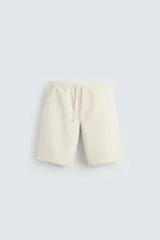 Bermuda color beige de denim de algodón, corte relaxed fit, con cintura elástica ajustable con cordón, bolsillos frontales y traseros de vivo.