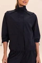 Campera negra de corte ancho y mangas raglán, confeccionada en tejido ligero y transpirable resistente al agua. Cuenta con capucha alta con cierre de cremallera y botones, mangas y barra ajustables, y bolsillo funcional en la parte delantera.