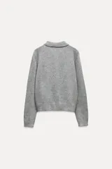 Chaqueta de punto gris con cuello solapa y manga larga. Tiene bolsillos delanteros de plastrón, rib en los acabados y cierre frontal con botones a presión en forma de lazo.