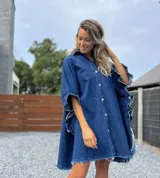Camisola tipo poncho en jean azul oscuro con cuello camisero, cierre frontal con botones, mangas cortas amplias con volados y ruedo con flecos.