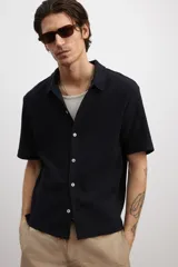 Camisa negra de punto con textura, de corte recto, manga corta, cuello inglés, tapeta lisa, canesú en la espalda y bajo recto con aberturas laterales.