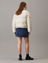 Campera puffer color beige claro con logo de Calvin Klein bordado en el pecho. Tiene cuello alto, cierre con cremallera y botones a presión, bolsillos laterales y puños elásticos.