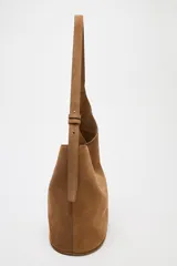Bolso tipo saca asimétrica de serraje color marrón. Tiene asa bandolera ajustable y cierre con imán. Incluye cartera con bolsillo extraíble.