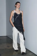 Vestido satinado negro con escote pico y tirantes finos. Presenta aplicación de encaje en el escote y en el bajo asimétrico, que incluye un tajo o caída pronunciada.