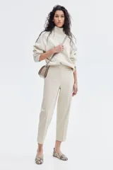 Pantalón cigarette color beige de tiro alto, con cierre y corchetes ocultos, bolsillos laterales discretos y bolsillos insertados decorativos atrás. Piernas ajustadas con abertura en los bajos y pliegues adelante y atrás.