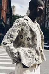 Blazer de tejido con estructura color crudo, con estampado de flores bordadas en negro. Tiene cuello solapa, manga larga y cierre frontal con botones negros.